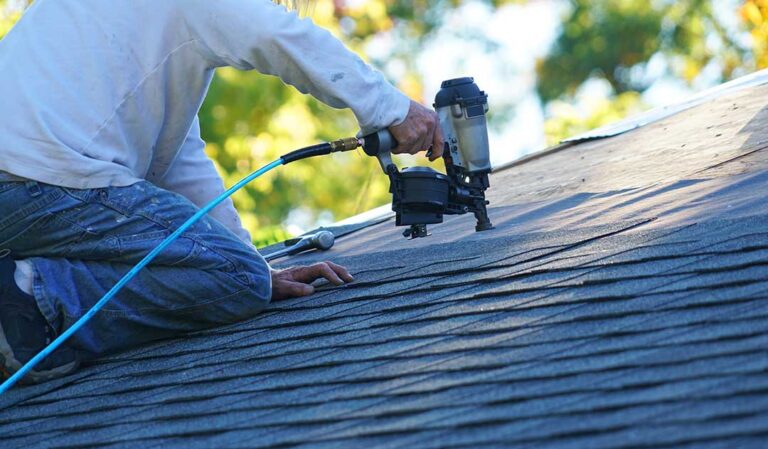 A homeowner uses a ladder and plastic roof rake to remove leaves from a shingle roof.