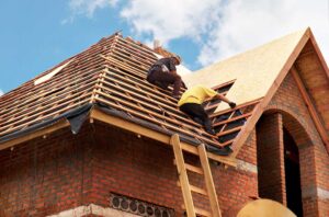 Person inspecting a leaf-covered roof and clearing debris from a gutter beside a residential home.