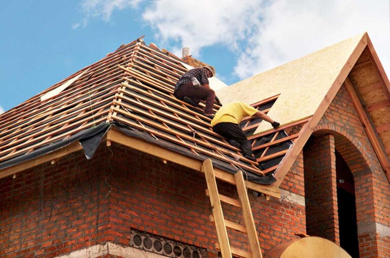 Person inspecting a leaf-covered roof and clearing debris from a gutter beside a residential home.